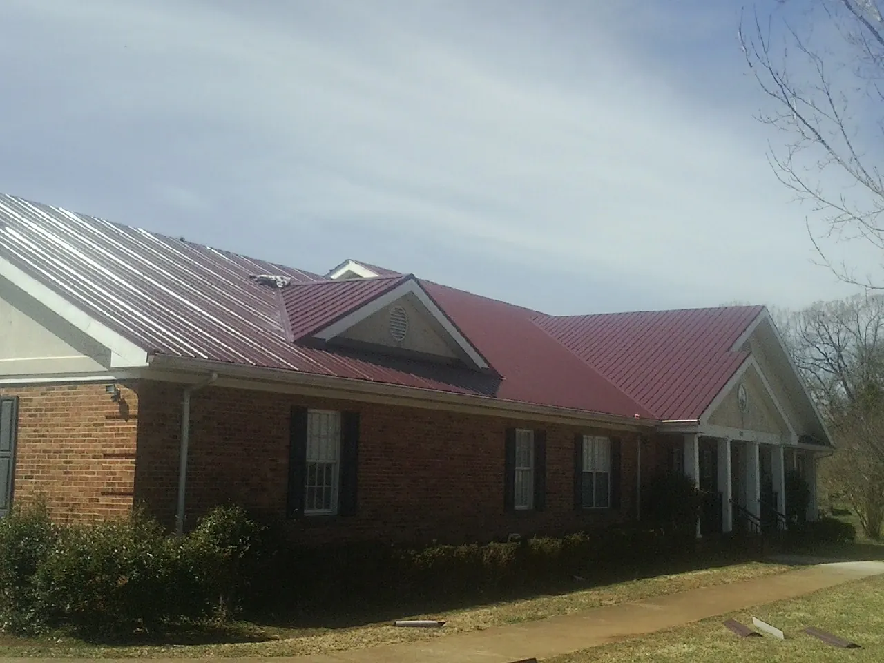 Red metal roof with gabled entries on a long brick building, from a Hail Damage Roof Repair job in College Point