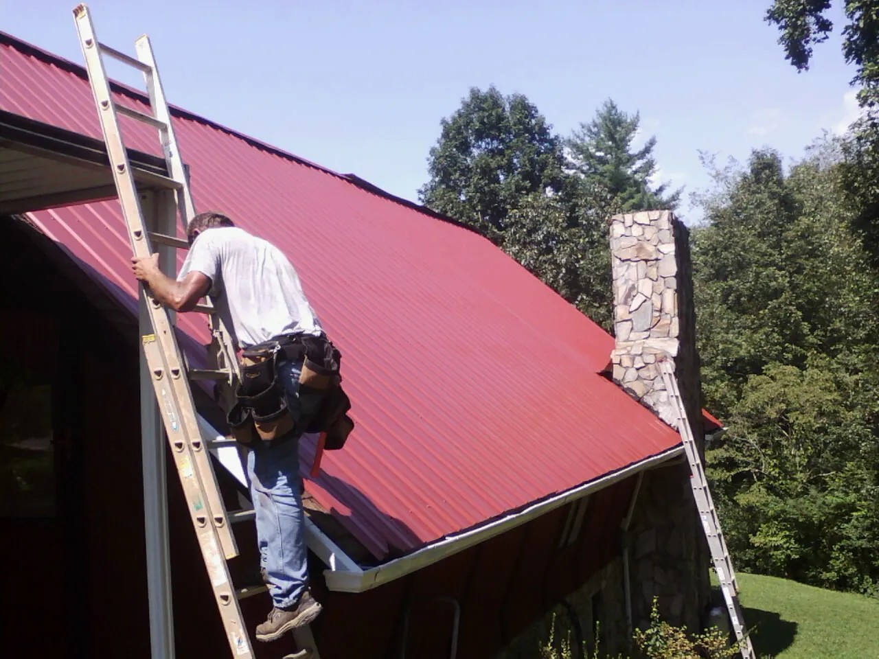 Roofer on a ladder beside a red metal roof and stone chimney — Soffit & Fascia Repair by Roofing Brothers in College Point