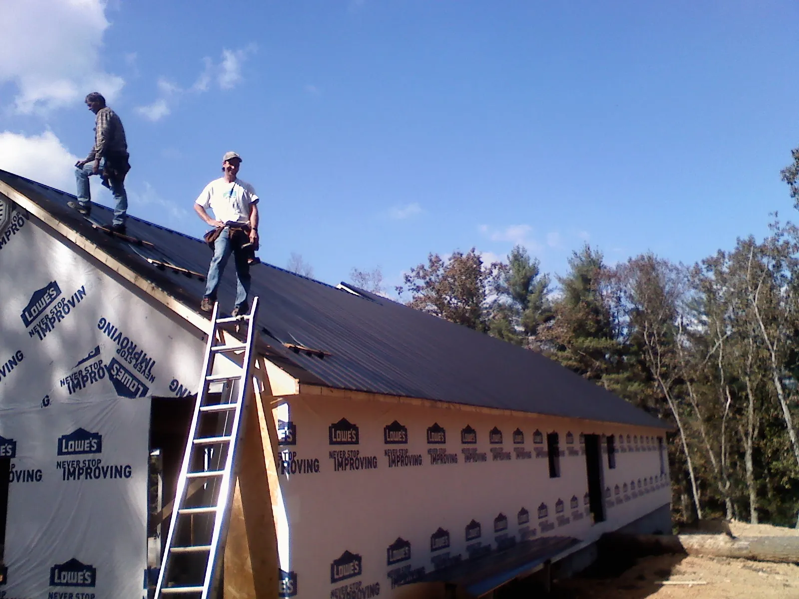 Two roofers standing on a long dark metal roof over a wrapped building in College Point